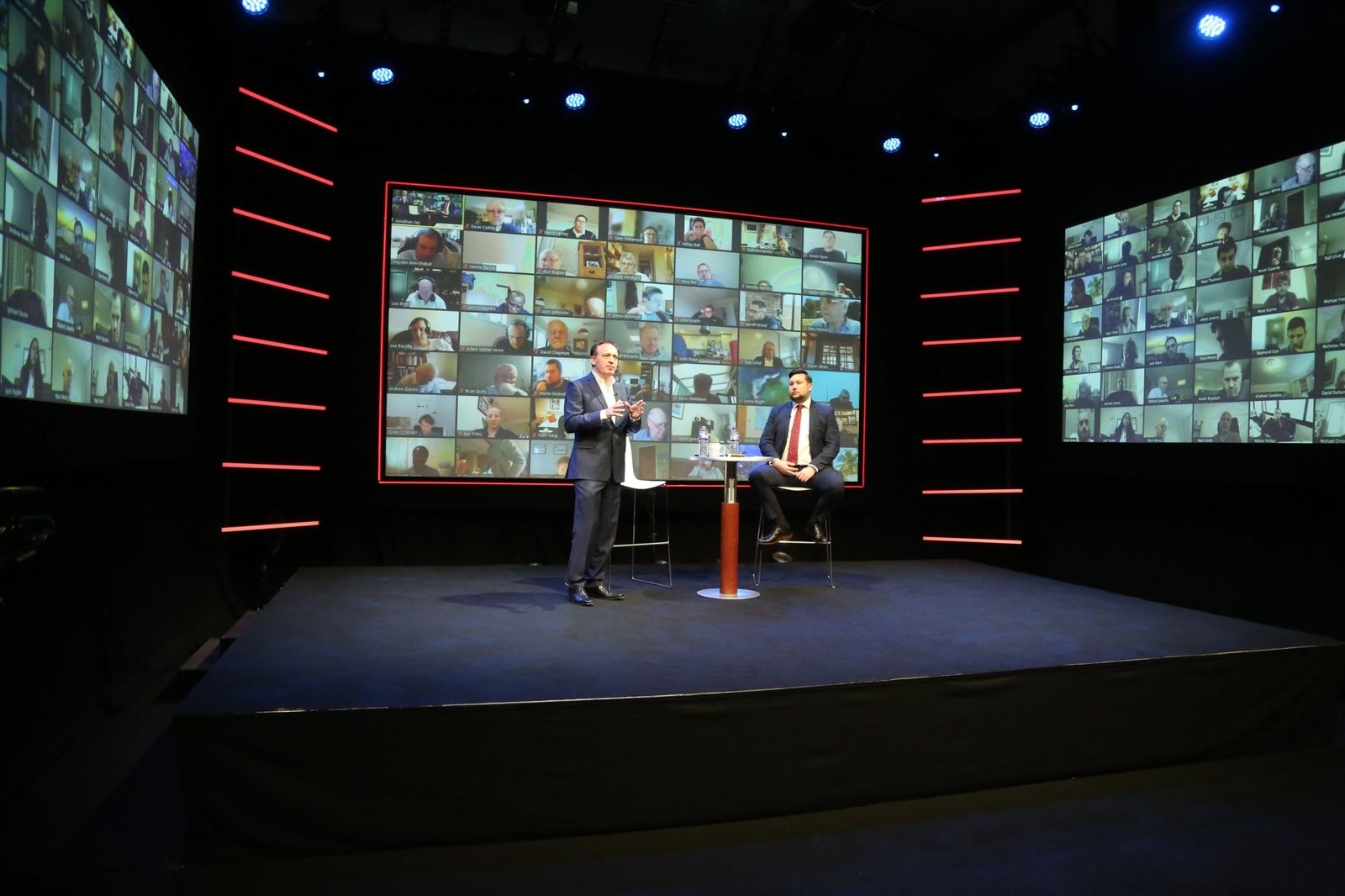 Two presenters in suits on stage, with the remote audience on video walls